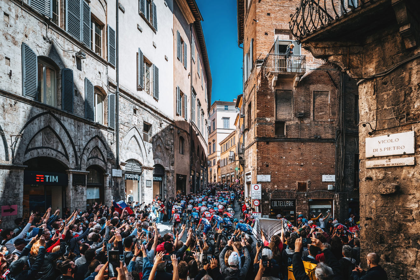 Зрители на узких улочках Сиены, Италия, наблюдают за прохождением группы велосипедистов